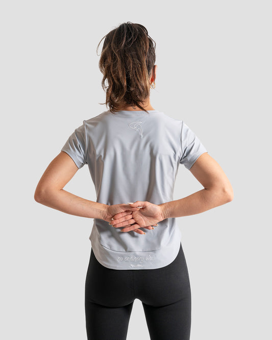 GoofyPRO Cayton Core Performance Tshirt in cloud grey with short sleeves, rear. White embroidered 'no ordinary way' in white on waistband. White embroidered logo at top of neck. worn by female model